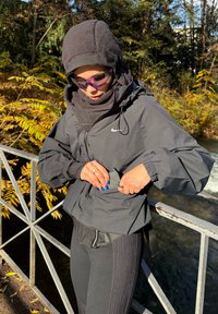 Mustast hõlmikuga kerge jope kapuutsiga ja pöidlahõlmadega, kihiliselt üle tumeda pluusi ja soonikpükstega, kombineeritud värviliste päikeseprillidega.