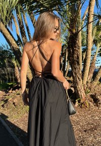 Sort ryggløs kjole med spaghettistropper, rynkedetaljer ved midjen og et flytende skjørt. Brukt med en liten svart veske foran palmetrær.