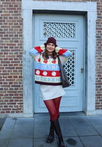 Jeune femme posant en souriant, portant un pull coloré avec l'inscription "Tuesday Weekend", des collants bordeaux, des bottes et un bonnet devant une porte ornée bleue.