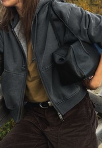 Szara bluza z kapturem zapinana na zamek, z teksturowaną podszewką, nałożona na brązową koszulę, w połączeniu z ciemnymi sztruksowymi spodniami i czarną skórzaną torbą.