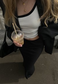 Crna duga suknja, bijela rebrasta crop majica, crni blejzer, zlatni lančići, drži hladno piće s prugastom slamkom, crna torba vidljiva.
