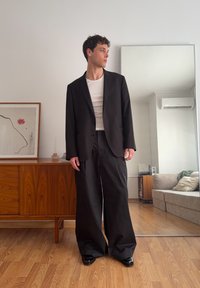 Jeune homme aux cheveux courts et bouclés portant un pantalon noir ample, un blazer noir, une chemise blanche, debout dans un salon avec un buffet en bois et un grand miroir.