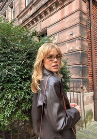 Braune Lederjacke mit glatter Textur, schulterlange blonde Haare und hellbraune Katzenaugenbrille, vor grünem Laub und Backsteinarchitektur.