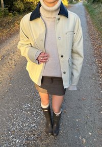 Jasnopopielaty płaszcz z ciemnym kołnierzem, w połączeniu z kremowym golfem i szarym spódnicą. Czarne gumowe kalosze z szarymi skarpetami. Casualowe otoczenie na zewnątrz.