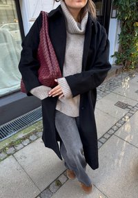 Čierny dlhý kabát cez svetlosivý rolák, doplnený sivými nohavicami širokého strihu a hnedými členkovými čižmami. Nosená je bordová tkaná taška.