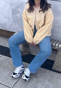 Személy bézs színű polárdzsekit, kék farmert és fehér Nike sportcipőt viselve egy fém padon ül, mögötte kőfal.