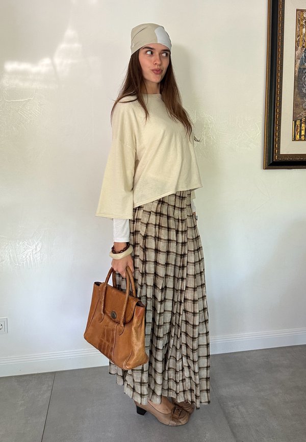 Mujer con top beige, falda larga a cuadros, pañuelo en la cabeza y tacones sostiene un bolso de cuero marrón mientras está de pie contra una pared blanca con arte enmarcado.