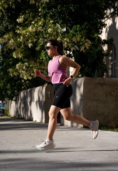 Atleta con una camiseta sin mangas rosa y pantalones cortos negros, corriendo por una acera; lleva gafas de sol y zapatillas de correr ligeras con una suela texturizada.