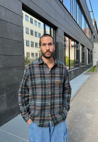 Homem com barba vestido com uma camisa xadrez preta e cinza e jeans azul está de pé do lado de fora, perto de um edifício moderno com grandes janelas.