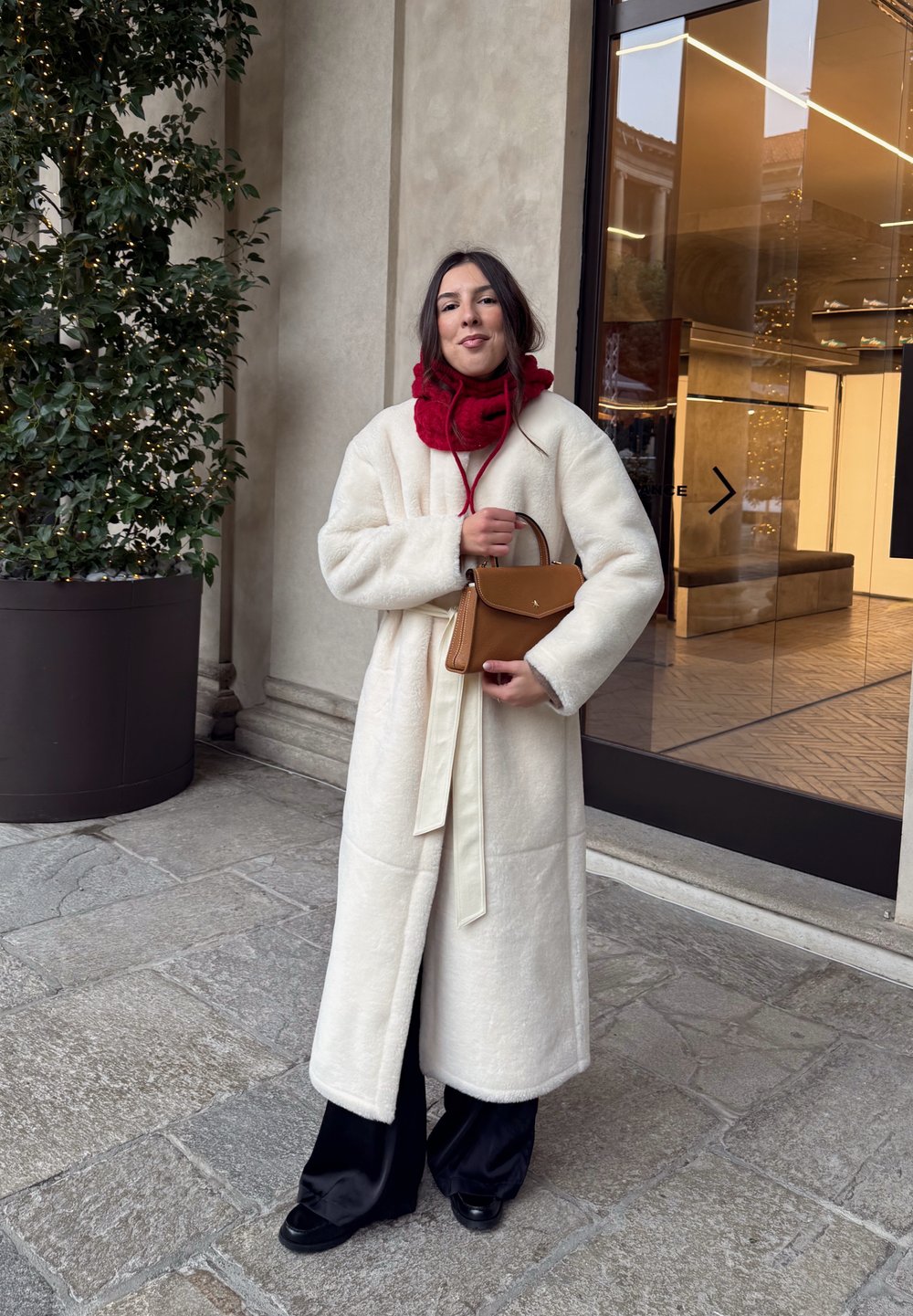 Lang cremefarvet frakke med blød tekstur og bælte, kombineret med et rødt tørklæde. Holder en lille brun taske. Sorte bukser med vide ben og sorte sko.