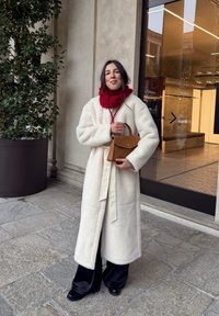 Lang cremefarvet frakke med blød tekstur og bælte, kombineret med et rødt tørklæde. Holder en lille brun taske. Sorte bukser med vide ben og sorte sko.