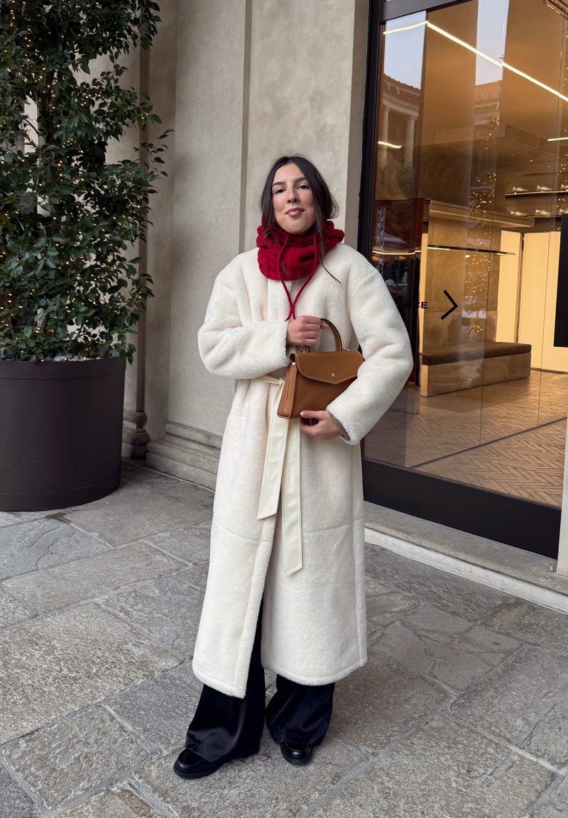 Pikk kreemjas mantel pehme tekstuuri ja vööl, kombineeritud punase salli. Kätel on väike pruun kott. Mustad laiade säärtega püksid ja mustad jalanõud.