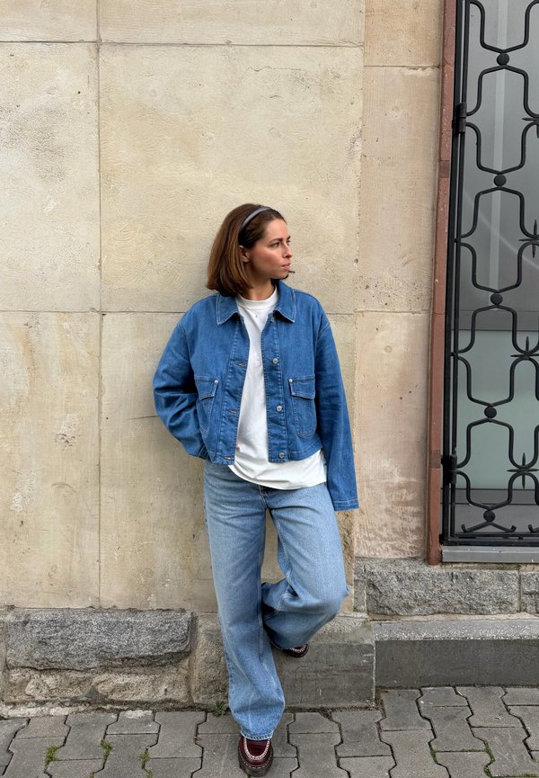 Una mujer con una chaqueta vaquera y vaqueros se apoya en un muro de piedra junto a una puerta metálica decorativa.