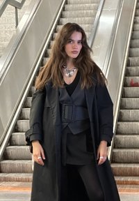 Palton negru peste o ținută stratificată, având o curea texturată și un colier din argint. Părul este ondulat. Fundalul prezintă scări rulante.