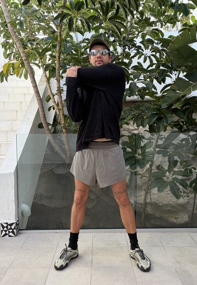 Hombre con gafas de sol, camiseta negra de manga larga y pantalones cortos grises estira el brazo cruzado sobre el pecho en un patio exterior con plantas detrás de él.