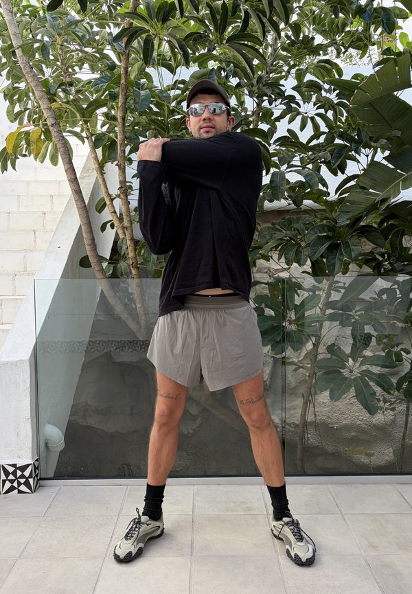 Man wearing sunglasses, black long-sleeve shirt, and grey shorts stretches arm across chest on outdoor patio with plants behind him.