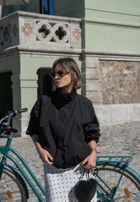Čierna ľahká bunda s vysokým golierom, skombinovaná s bielou sukňou s bodkami a okrúhlou čiernou kabelkou. Vedľa bicykel s tyrkysovým rámom.