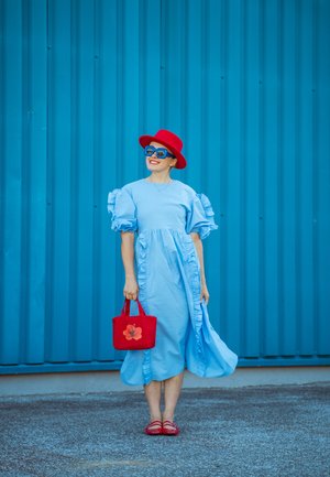 Vestido azul claro con volantes y mangas cortas; sombrero rojo y gafas de sol. Sostiene un bolso rojo con un diseño floral; sandalias rojas. Fondo azul.