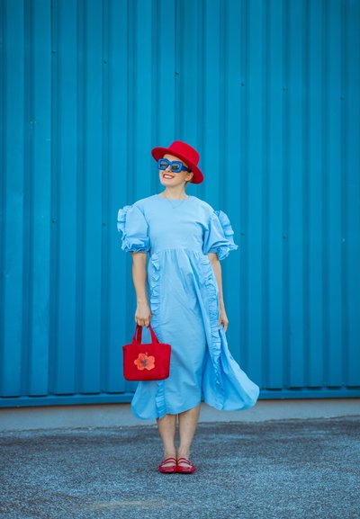 Vestido azul claro con volantes y mangas cortas; sombrero rojo y gafas de sol. Sostiene un bolso rojo con un diseño floral; sandalias rojas. Fondo azul.
