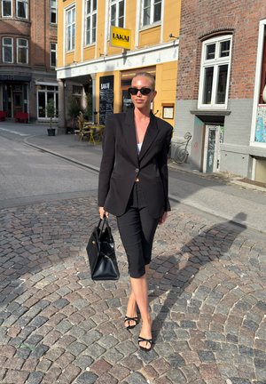 Schwarze maßgeschneiderte Blazer über einem weißen Oberteil, kombiniert mit schwarzen knielangen Hosen. Schwarze Handtasche und Riemchenabsätze; Hintergrund mit Kopfsteinpflasterstraße.