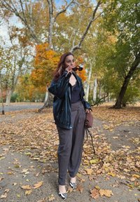 Melna ādas jaka, tumša blūze, pelēkas bikses un svītrainas zebrās raksta augstpapēžu kurpes. Tur rokās saulesbrilles, stāv uz lapām klāta ceļa ar kokiem apkārt.