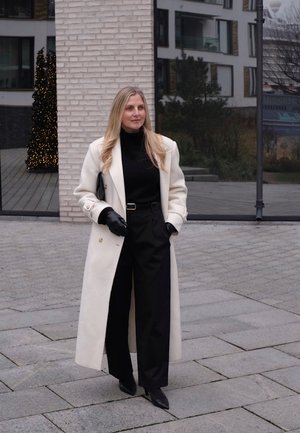 Abrigo largo color crema sobre un cuello alto negro, combinado con pantalones anchos negros. Los accesorios incluyen guantes negros y un bolso de mano. Texturas lisas y confeccionadas a medida.