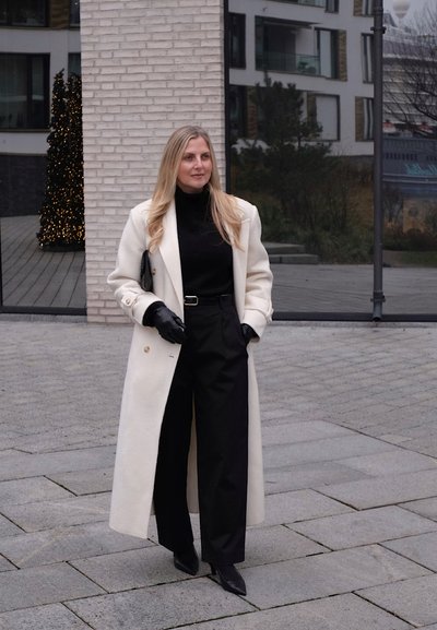 Abrigo largo color crema sobre un cuello alto negro, combinado con pantalones anchos negros. Los accesorios incluyen guantes negros y un bolso de mano. Texturas lisas y confeccionadas a medida.