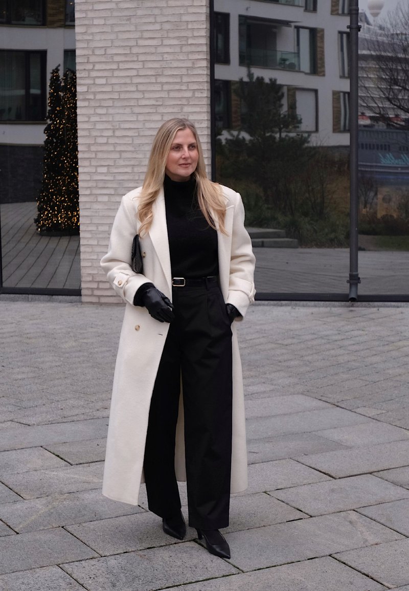 Lungo cappotto color crema sopra un dolcevita nero, abbinato a pantaloni neri a gamba larga. Accessori comprendono guanti neri e una pochette. Texture lisce e sartoriali.