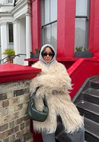 Personne portant un manteau shaggy beige, un sweat à capuche gris et des lunettes de soleil noires, appuyée contre un mur en briques avec une rampe rouge près des escaliers d'un bâtiment.