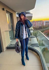 Chaqueta de cuero negro, camisa blanca, leggings azul marino, botines teal. Sosteniendo un bolso oscuro, de pie en un balcón con barandilla geométrica.