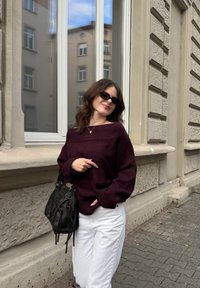 Preveliki bordo džemper s dizajnom koji otkriva ramena, u kombinaciji s bijelim širokim hlačama. Crna teksturirana torba s remenima i ogrlica s privjeskom.