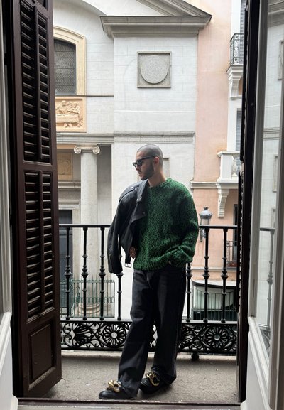 Suéter verde texturizado, chaqueta de cuero negra, pantalones de mezclilla oscuros y zapatos negros con acentos dorados, todo con una vista decorativa de un balcón detrás.