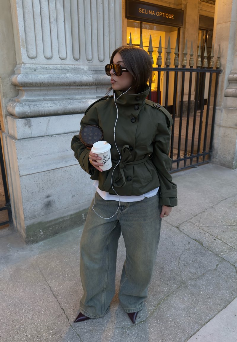Frau in übergroßer grüner Jacke, weiten Jeans, Sonnenbrille, hält Kaffeetasse und runde Tasche, steht neben einem Steingebäude mit Eisentor.