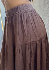 Brązowa, teksturowana spódnica z wysokim stanem noszona przez osobę stojącą przy jasnej, teksturowanej ścianie, ukazująca okolicę talii i bioder.