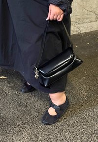 Černá, lesklá kabelka se štíhlým designem, která má stříbrné srdíčko jako přívěsek a krátký ramenní popruh, doplněná černou obuví.