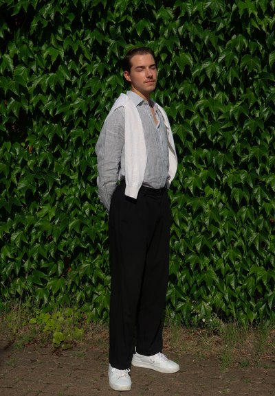 Una persona está de pie contra un fondo de hiedra verde, vistiendo una camisa a rayas, pantalones negros y zapatillas blancas, con un suéter blanco sobre los hombros.