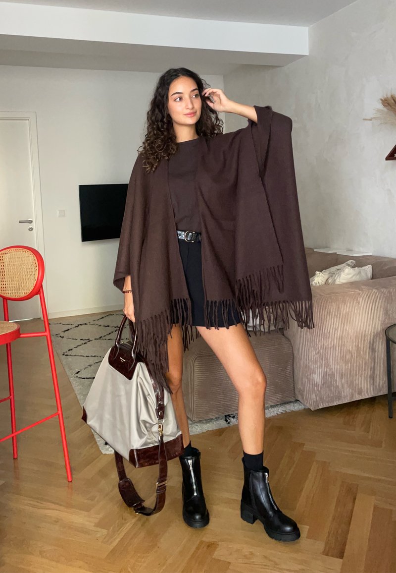 Femme avec des cheveux bouclés portant un châle frangé marron, un short noir et des bottes, tenant un grand sac beige et marron dans un salon moderne.