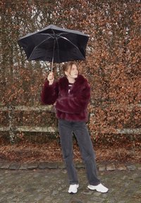 Femme en manteau de fourrure bordeaux et pantalon foncé tenant un parapluie noir, debout sur un chemin pavé, avec des feuilles d'automne et des arbres nus en arrière-plan.