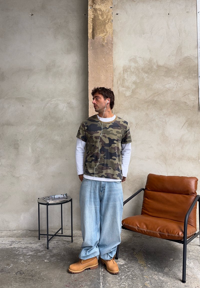 Camo-muster T-Shirt, darunter weisses langärmeliges Shirt, hellblaue weite Jeans, beigefarbene Arbeitsstiefel. Brauner Leder-Sessel neben einem schwarzen Beistelltisch.