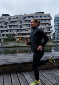 Fekete steppelt dzseki, fekete leggings és lime zöld futócipők. Fa sétány és épületek a háttérben, borús ég alatt.