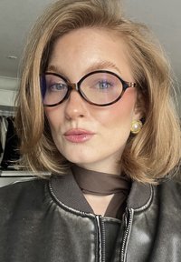 Mustat nahkainen bomber-takki, jossa on vetoketju edessä, pyöreät kilpikonnalasi, ja kuvioitu ruskea huivi. Kulta-sävyiset korvakorut, joissa on helmikoriste.