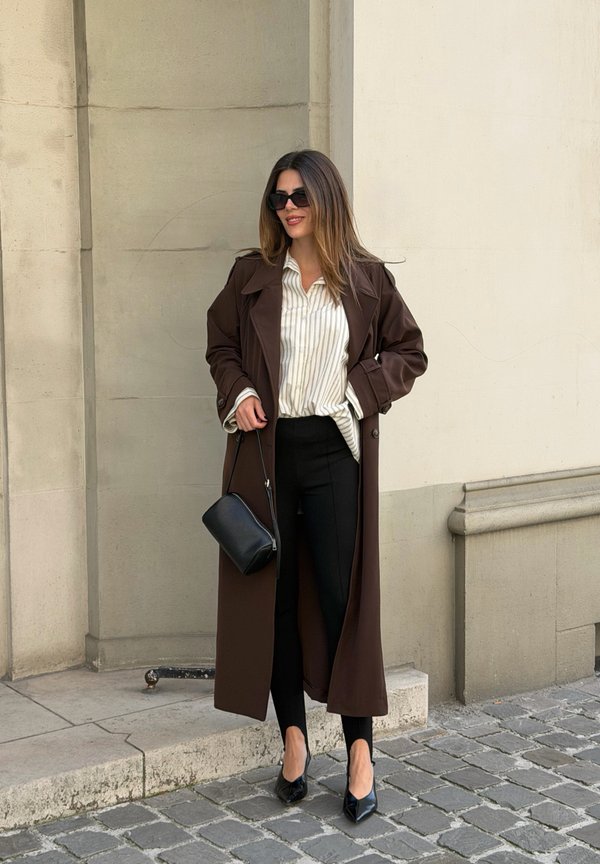 Mujer con un abrigo marrón largo, camisa de rayas, pantalones negros, tacones negros y gafas de sol, de pie en una acera empedrada junto a una pared beige.