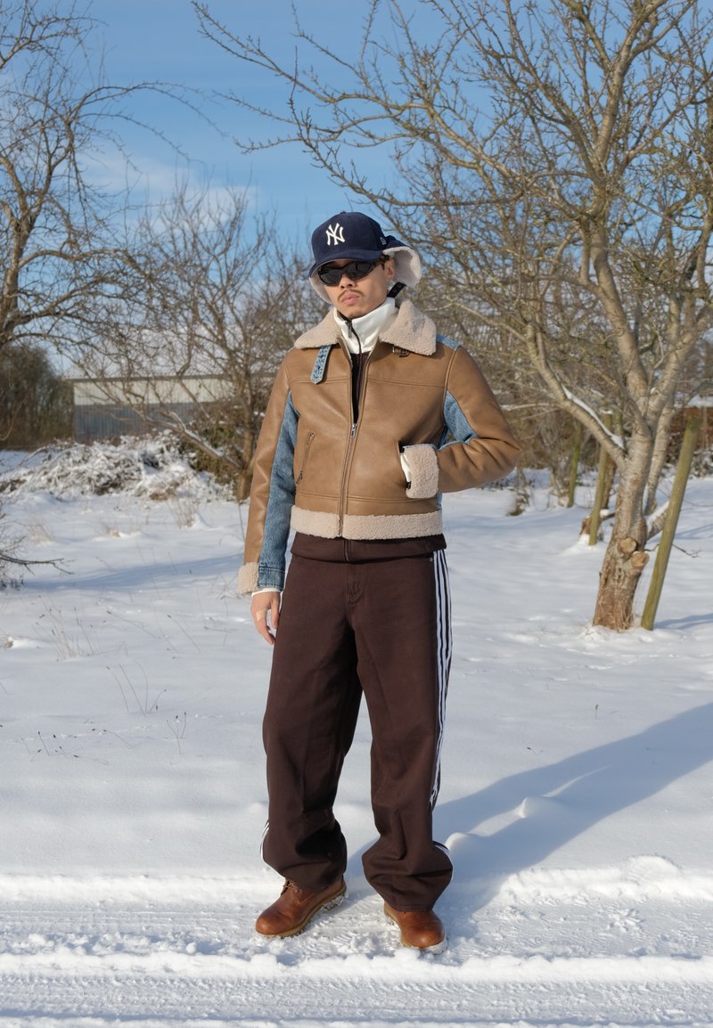 Man som bär en brun fårskinnsjacka, svarta byxor med vita ränder, bruna stövlar, solglasögon och keps står på en snötäckt stig med kala träd.