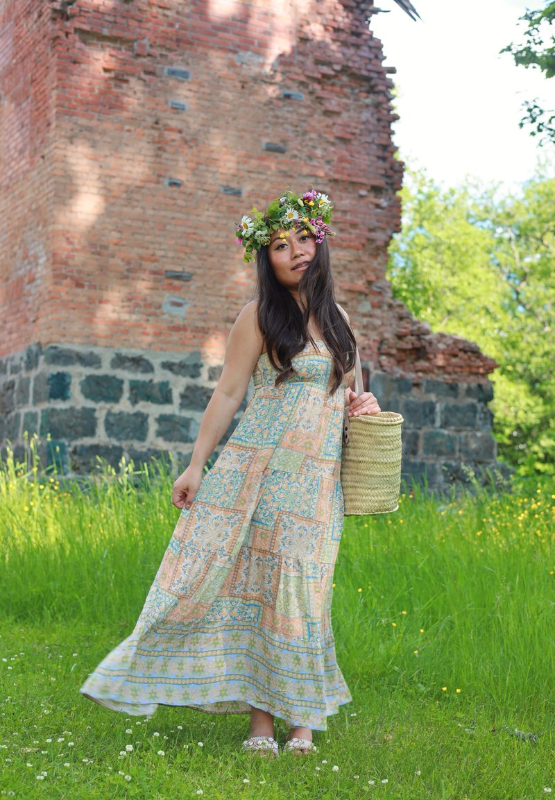Woman in a floral crown and patterned dress holding a wicker basket stands on green grass by an old brick and stone wall in sunlight.