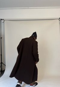 Muž v dlouhém tmavém kabátě, černé čepici a kalhotách s potiskem prochází kolem jednoduchého bílého pozadí fotografického studia s podpěrnými stojany.