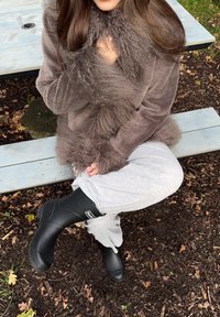 Ruda dirbtinio kailio paltas, pilkos sportinės kelnės ir juodi guminiai kulkšnių aulinukai. Sėdi ant medinės suolelio, su natūralia ir kaimiška dirvos bei lapų fone.