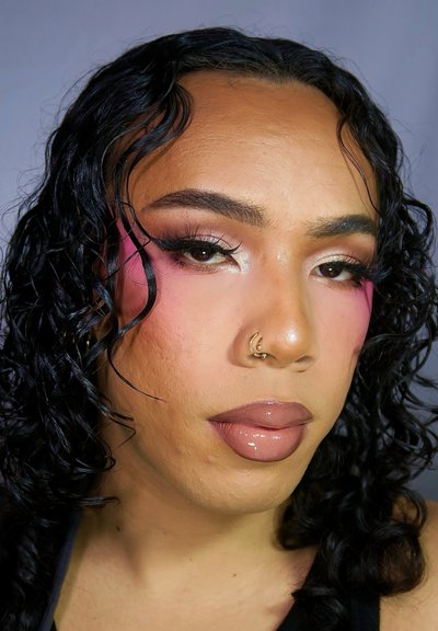 Primer plano de una persona con cabello rizado y negro, que lleva rubor rosa, delineador de ojos intenso y un brillo labial nude, con un aro dorado en la nariz.