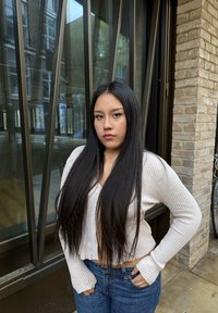 Lange schwarze Haare, trägt einen cropped, gerippten cremefarbenen Pullover und eine blaue Jeans, steht vor einer Glaswand mit metallischen Akzenten.