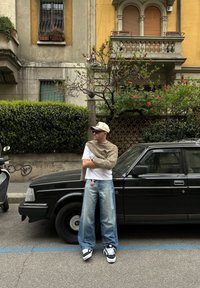 Svart bil parkert på gaten. Individ som har på seg en hvit skjorte, overdimensjonerte lyseblå jeans og en lys strikkegenser, samt mørke solbriller og en caps.