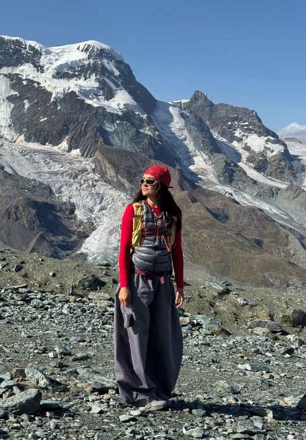 Une randonneuse portant un bandana rouge et un équipement de plein air se tient sur un terrain rocheux, devant des montagnes enneigées et des glaciers.